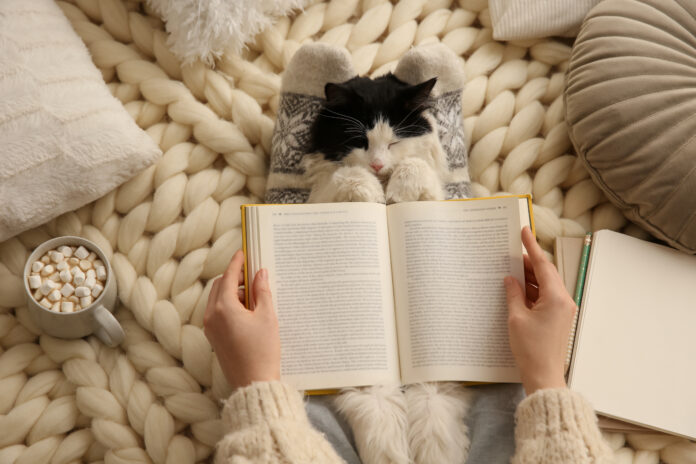 Woman,Reading,Book,And,Holding,Adorable,Cat,On,Knitted,Blanket,